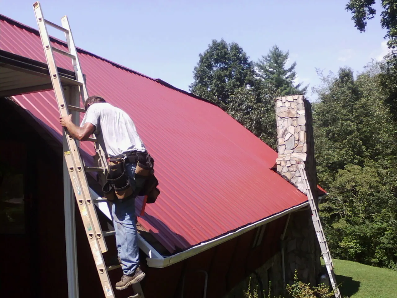 Roofer on a ladder beside a red metal roof and stone chimney — Soffit & Fascia Repair by Roofing Brothers in Newark